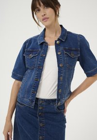 Denim jasje met korte mouwen, lichtblauwe tint, twee borstzakken, metalen knopen en versleten accenten. Gecombineerd met een denim rok.
