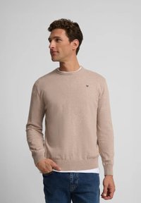 Suéter de punto beige con cuello redondo, puños y cintura de canalé. Pequeño logo bordado en el lado izquierdo del pecho. Combinado con unos vaqueros azules.