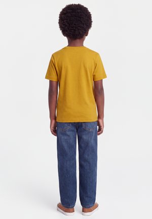 Enfant aux cheveux bouclés portant un t-shirt jaune moutarde à manches courtes, un jean bleu et des chaussures marron, debout de dos sur un fond uni.