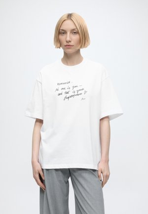 Jeune femme aux cheveux blonds courts portant un t-shirt blanc avec un texte manuscrit, associé à un pantalon gris clair, debout devant un fond blanc.