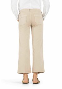 Beige weit geschnittene Hose aus Baumwollmischung mit hohem Bund, zwei Gesäßtaschen und einem sauberen Saum. Getragen mit flachen Sandalen.