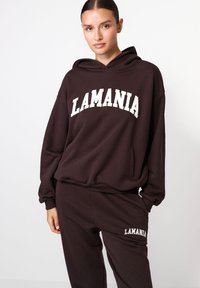 Hnedá mikina s kapucňou a bielym textom "LAMANIA" cez hruď, s klokaním vreckom a rebrovaným lemom. V páre s rovnakými teplákmi.