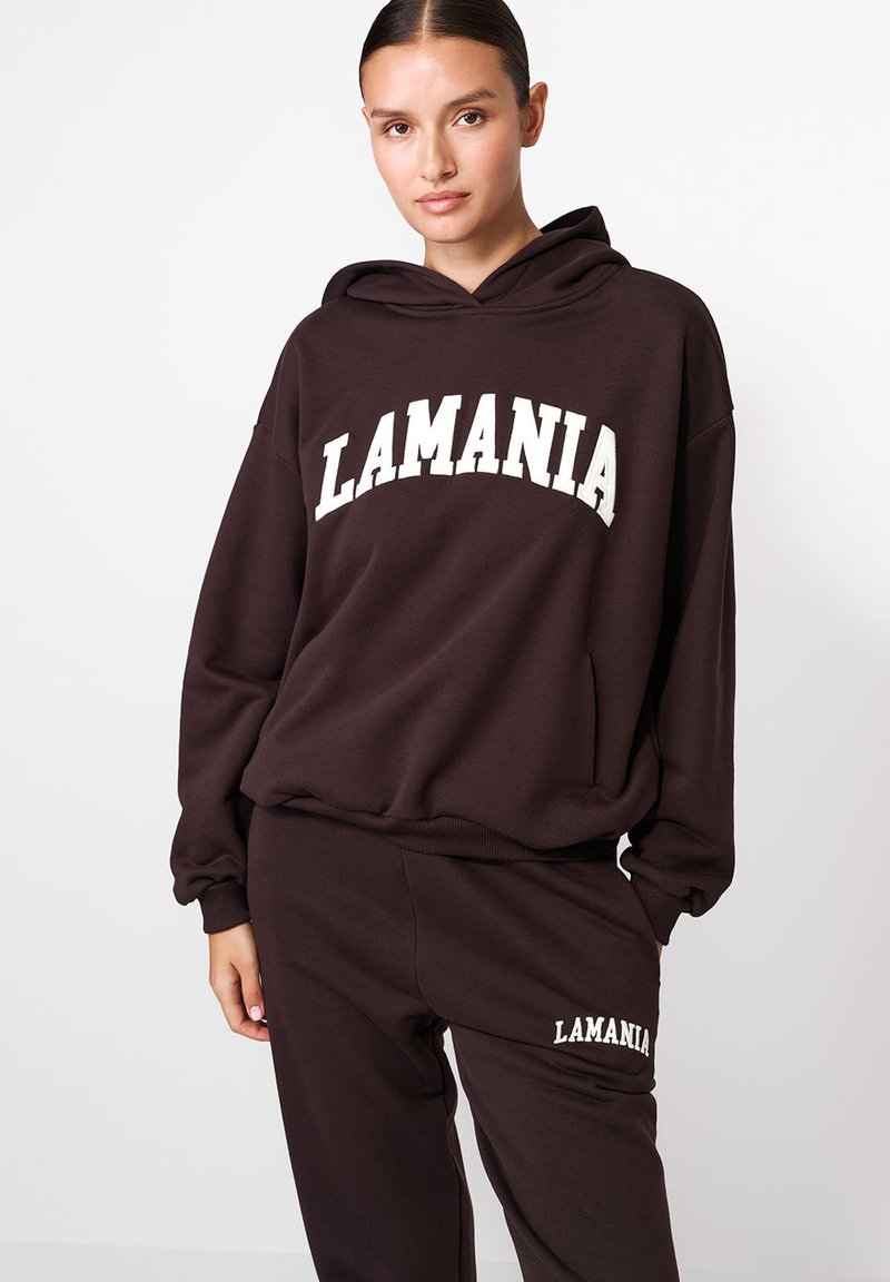 Hnedá mikina s kapucňou a bielym textom "LAMANIA" cez hruď, s klokaním vreckom a rebrovaným lemom. V páre s rovnakými teplákmi.