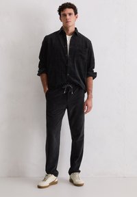 Mann in einem schwarzen Hemd, schwarzen Jogginghosen und beigen Sneakers, der gegen eine weiße strukturierte Wand steht.