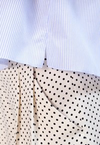 Světle modrá pruhovaná košile s rozparkem, která je vrstvená přes krémovou sukni s černými puntíky, zdůrazňující texturu a kontrast.