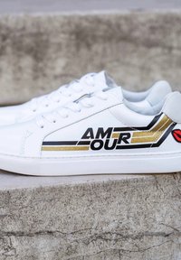 Baskets blanches avec une finition lisse, un design à rayures noires et dorées, et le mot "AMOUR" en lettres grasses, avec une touche de rouge.