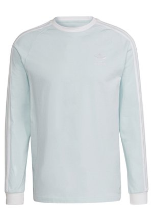 Mies, joka pukeutunut vaaleansiniseen pitkähihaseen Adidas-paitaan, jossa valkoiset raidat hihoissa, ja harmaisiin Adidasin cargohousuihin, käsi taskussa.