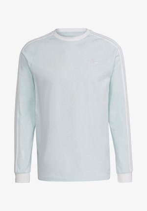 Mies, joka pukeutunut vaaleansiniseen pitkähihaseen Adidas-paitaan, jossa valkoiset raidat hihoissa, ja harmaisiin Adidasin cargohousuihin, käsi taskussa.