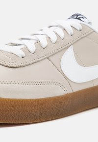 Beige sneaker med vit Nike swoosh, tillverkad av läder och mocka, med en texturerad gummisula och vita snörning.