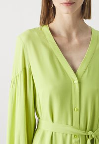 Lime green button-up dress with long sleeves and a v-neckline. Soft texture, cinched waist with a matching belt, and subtle pleats at shoulders.