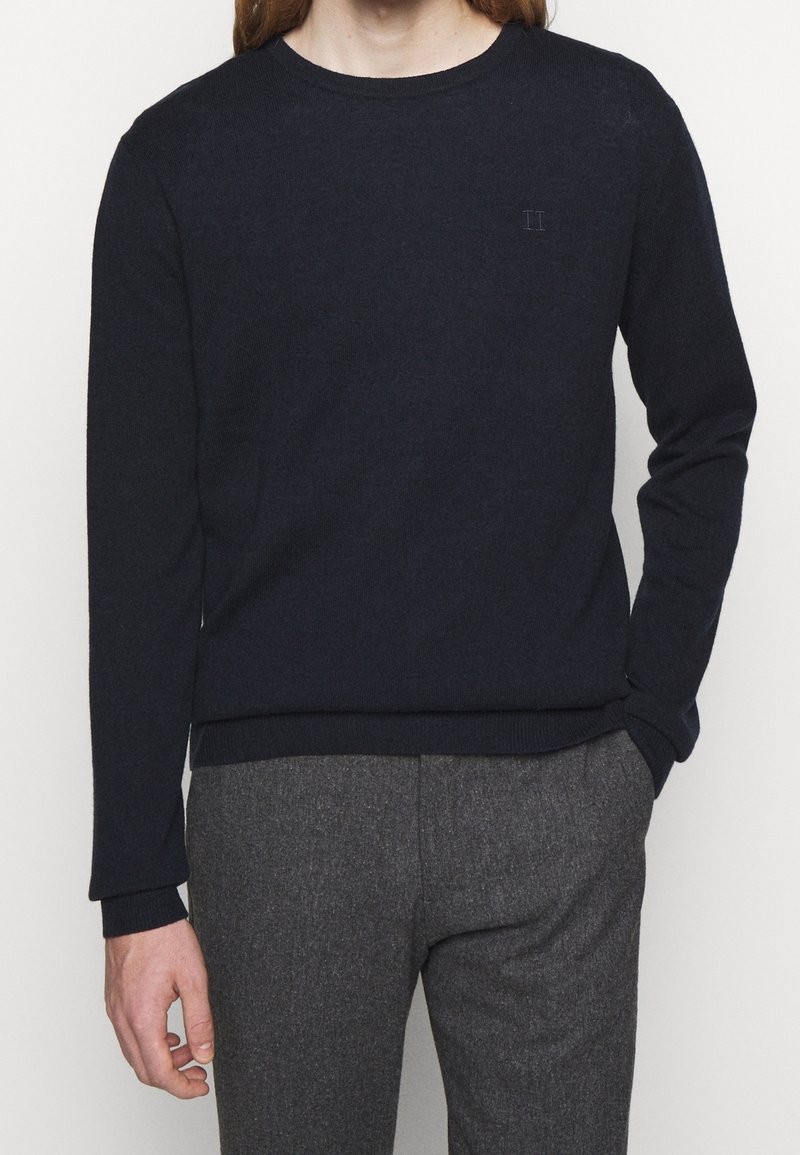 Marinblå stickad tröja med rund halsringning, långa ärmar och ett subtilt texturerat mönster. Har en liten, ton-i-ton logga på bröstet.