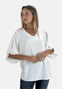 Witte, lichte blouse met V-hals en korte, wijde mouwen. Heeft een gladde textuur en een ontspannen pasvorm, gecombineerd met donkere jeans.