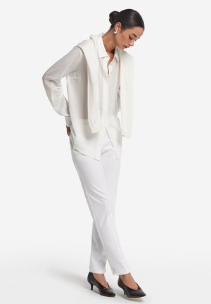 White button-up shirt, white pants, and a light sweater draped over shoulders. Black pointed heels. Smooth fabric, casual design.