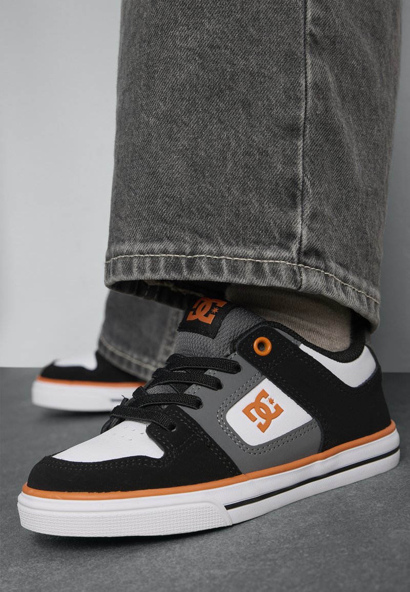 Čierne a šedé skateboardové topánky s oranžovými akcentmi, vybavené polstrovaným golierom, bielou podrážkou a detailmi loga. Viditeľný textúrovaný materiál a šnúrky.