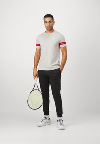 Lys grå kortermet t-skjorte med to røde striper, svarte bukser og hvite joggesko, holder en tennisracket, står på en grå bakgrunn.