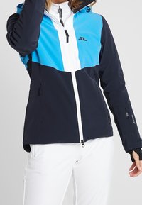 Modrá a námořnicky modrá bunda s barevnými bloky, vyrobená z voděodolného materiálu, se zipem vpředu, dvěma bočními kapsami a kapucí. Kontrastní bílé detaily na zipu.