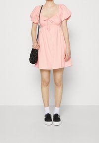 Vestido corto rosa con mangas abullonadas, escote cuadrado y detalle de lazo. Combinado con un bolso negro, calcetines blancos y zapatos negros tipo slip-on.