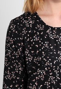 Black blouse with a floral pattern in pink and white. Features a round neckline and soft, textured fabric.