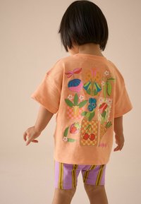 Barnens kortärmade skjorta i mjuk orange bomull har en färgglad grafik med frukt och blommor på ryggen. Kombineras med shorts med lila ränder.