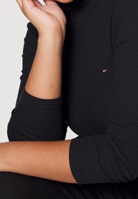 Sort turtleneck lavet af ribbet stof, med et lille mærke logo på brystet. Close-up af arme, der hviler mod kroppen.