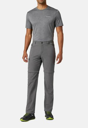 Mann trägt graues Columbia-T-Shirt mit kurzen Ärmeln, graue Zip-off-Cargohose und schwarze sowie grüne Sportschuhe, steht vor weißem Hintergrund.