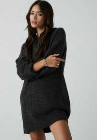 Czarna szara dzianinowa sukienka w oversize'owym fasonie z długimi rękawami, prążkowanym dołem i miękką fakturą. Modelka pozuje z założonymi rękami i bransoletką.