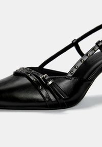 Čierne kožené slingback topánky s špicatou špičkou, ozdobené kovovými študmi a skríženým pásom. Hladká textúra s detailom s prackou.
