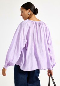Blusa oversize lilla in tessuto testurizzato, con colletto elasticizzato e maniche ampie. Presenta polsini con bottoni e una vestibilità rilassata.