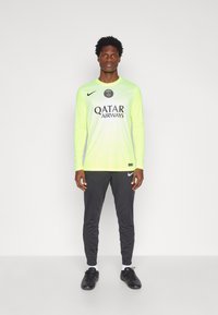 Garās piedurknes neona dzeltena sporta krekls ar melnu logotipu un tekstu; apvienots ar tumši pelēkiem šauriem biksēm un melniem futbola apaviem.