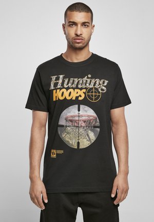 Melna kokvilnas T-kreklā ar dzeltenu un kamuflāžas tekstu "Hunting HOOPS". Centrālais attēls ir basketbola grozs mērķa rāmī.