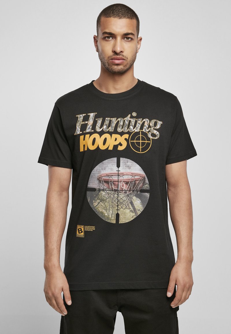 Juodas medvilninis marškinėliai su „Hunting HOOPS“ tekstu geltonai ir kamufliažo stiliaus. Viduryje esantis grafikas - krepšinio lankas taikinio rėmuose.
