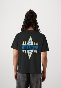 Mustat puuvillapaitat, joissa on suuri monivärinen graafinen kuva selkäpuolella. Suunnittelussa on sinistä tekstiä ja geometrisia muotoja kermanvärisellä taustalla.