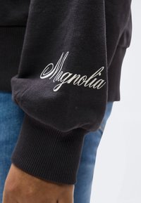 Μανίκι μαύρου φούτερ με λευκή καλλιγραφική επιγραφή «Magnolia», φορεμένο από άτομο με μπλε τζιν που φαίνεται στο φόντο.