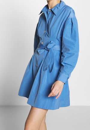 Vrouw met een blauwe jurk met knopen, lange opgeblazen mouwen en een gestrikte tailleband, staand tegen een effen lichte achtergrond.