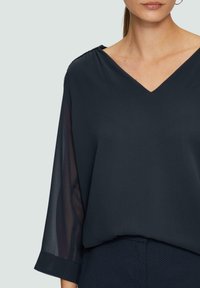 Dunkelbluse in Marineblau mit V-Ausschnitt und transparenten langen Ärmeln. Glatte Textur aus leichtem Stoff. Mit gerafften Details an den Schultern.