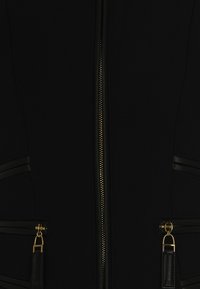 Chaqueta negra con una cremallera dorada, detalles en cuero y dos bolsillos con cremallera y herrajes dorados. Textura de tela suave con paneles laterales.