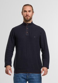 Dunkelblaues Pullover mit hohem Kragen, drei braunen Knöpfen und gerippten Bündchen. Strukturierter Stoff mit einem subtilen Muster und Logodetail.