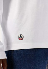 Camicia bianca a maniche lunghe con un piccolo logo ricamato circolare in blu e rosso sull'orlo. Il tessuto ha un aspetto liscio e strutturato.