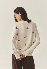Kremowy sweter z dzianiny z wielokolorową haftowaną dekoracją przedstawiającą wzory takie jak kwiaty, ptaki i postacie. Luźny krój z prążkowanymi mankietami i dekoltem.