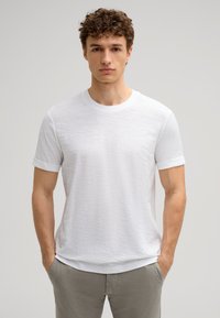 Weißes T-Shirt mit Rundhalsausschnitt, kurzen Ärmeln, texturiertem Stoff und lässigem Schnitt. Getragen mit hellgrauen Hosen und Händen in den Taschen.