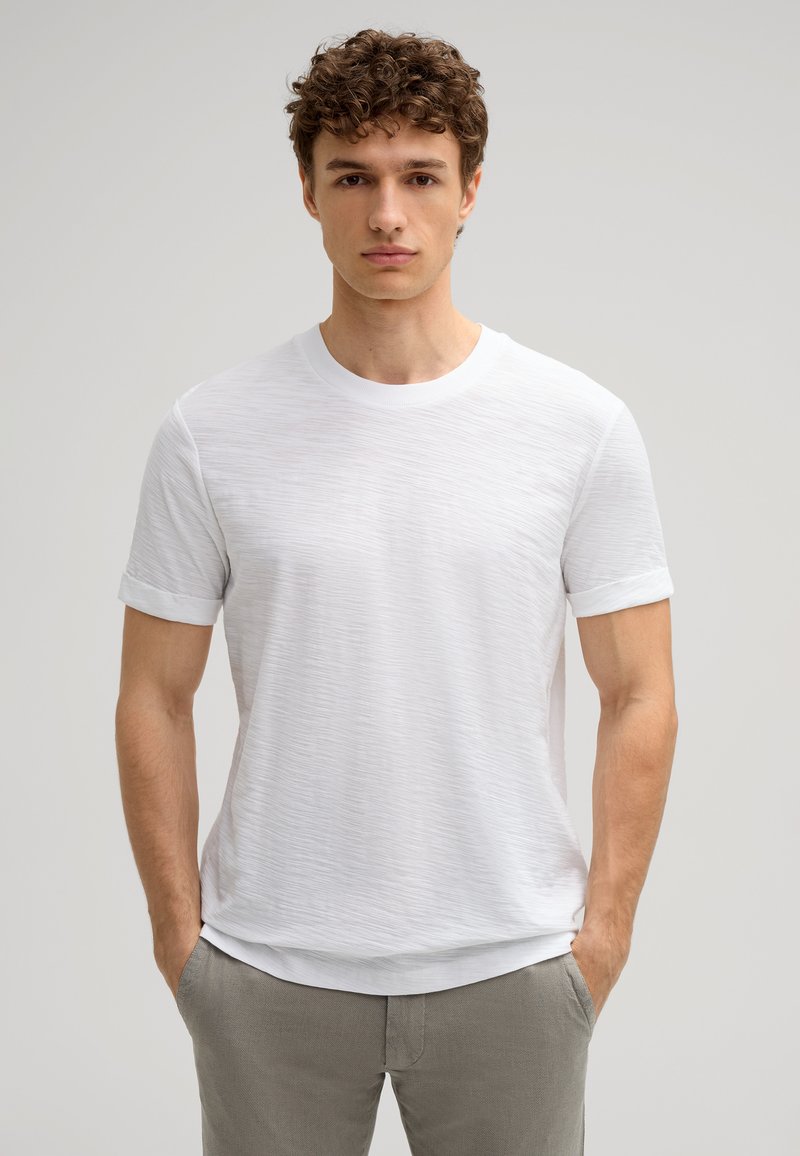 Weißes T-Shirt mit Rundhalsausschnitt, kurzen Ärmeln, texturiertem Stoff und lässigem Schnitt. Getragen mit hellgrauen Hosen und Händen in den Taschen.