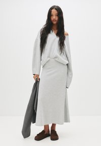 Sweat-shirt gris avec un design décolleté, associé à une jupe midi grise assortie. Le modèle tient une veste gris foncé. Des pantoufles marron complètent le look.