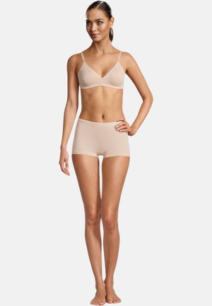 Frau steht mit einer Hand in der Hüfte, trägt einen BH in Nude-Ton und passende Boyshort-Unterwäsche vor einem schlichten weißen Hintergrund.