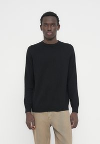 Pull noir en maille douce, avec un col rond et des manches longues. Porté avec un pantalon beige clair, sur un fond neutre.