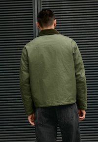 Grüne Bomberjacke mit strukturiertem Finish und einem Cordkragen, kombiniert mit dunklen Jeans. Der Hintergrund zeigt horizontale Latten.