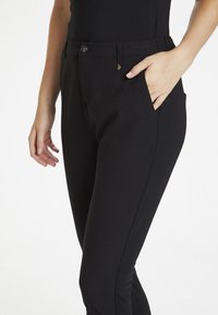 Pantalon noir en matériau extensible, avec une poche latérale, une fermeture à bouton et un logo discret près de la taille.