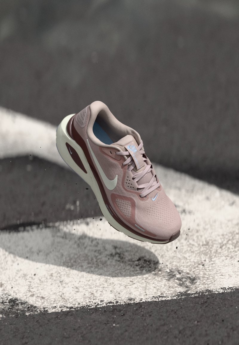 Ροζ ανοιχτό και μπορντό αθλητικό παπούτσι Nike που αιωρείται πάνω από άσφαλτο με λευκές βαμμένες γραμμές, εμφανίζοντας τη σόλα και τις πλευρικές λεπτομέρειες.