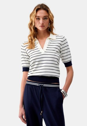 Donna con capelli biondi ondulati che indossa una maglietta polo bianca a maniche corte con righe blu navy e pantaloni blu navy con fascia in vita a righe, in piedi contro uno sfondo bianco.