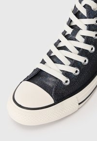 Adidași înălțați cu un material sclipitor de culoare albastru închis, șireturi albe și un capac al degetului din cauciuc alb, cu o talpă texturată. Accente din cauciuc negru.