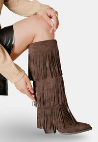 Bottes hautes en daim marron avec des franges, un bout pointu et un talon empilé, présentées aux côtés d'une main reposant sur la jambe.